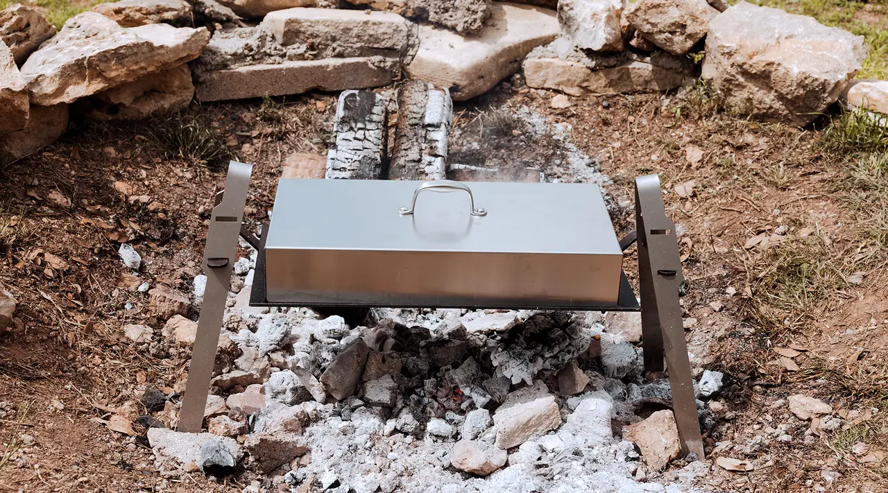 A metal box oven is set over a bed of hot coals from a campfire, situated outdoors surrounded by rocks and vegetation.