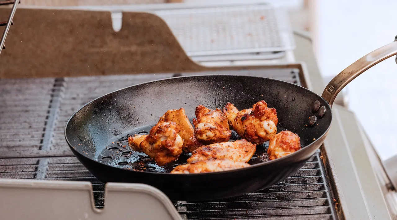 Seasoned chicken pieces are being cooked in a black frying pan on an outdoor grill.