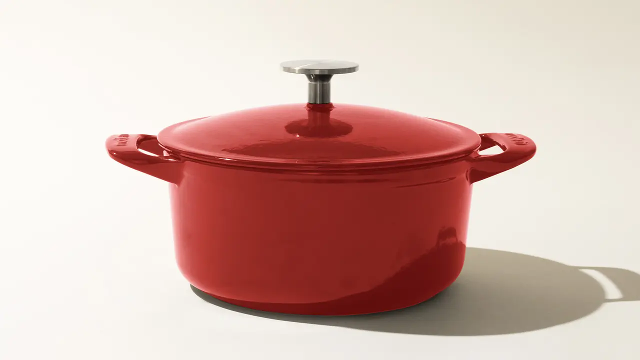 A shiny red enameled cast iron pot with a lid and stainless steel knob, positioned on a neutral background with a distinct shadow to the right side.