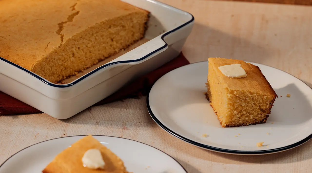 A freshly baked cornbread in a pan with a couple of slices served on small plates, one topped with a pat of butter.