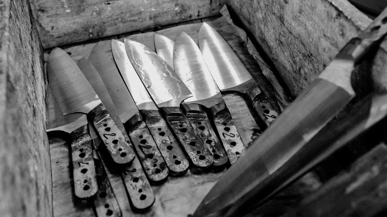 A collection of unfinished knives with wooden handles lying in a wooden box, shown in black and white.