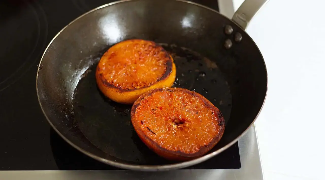 Two orange fruit halves with caramelized tops are being fried in a black skillet on a stovetop.