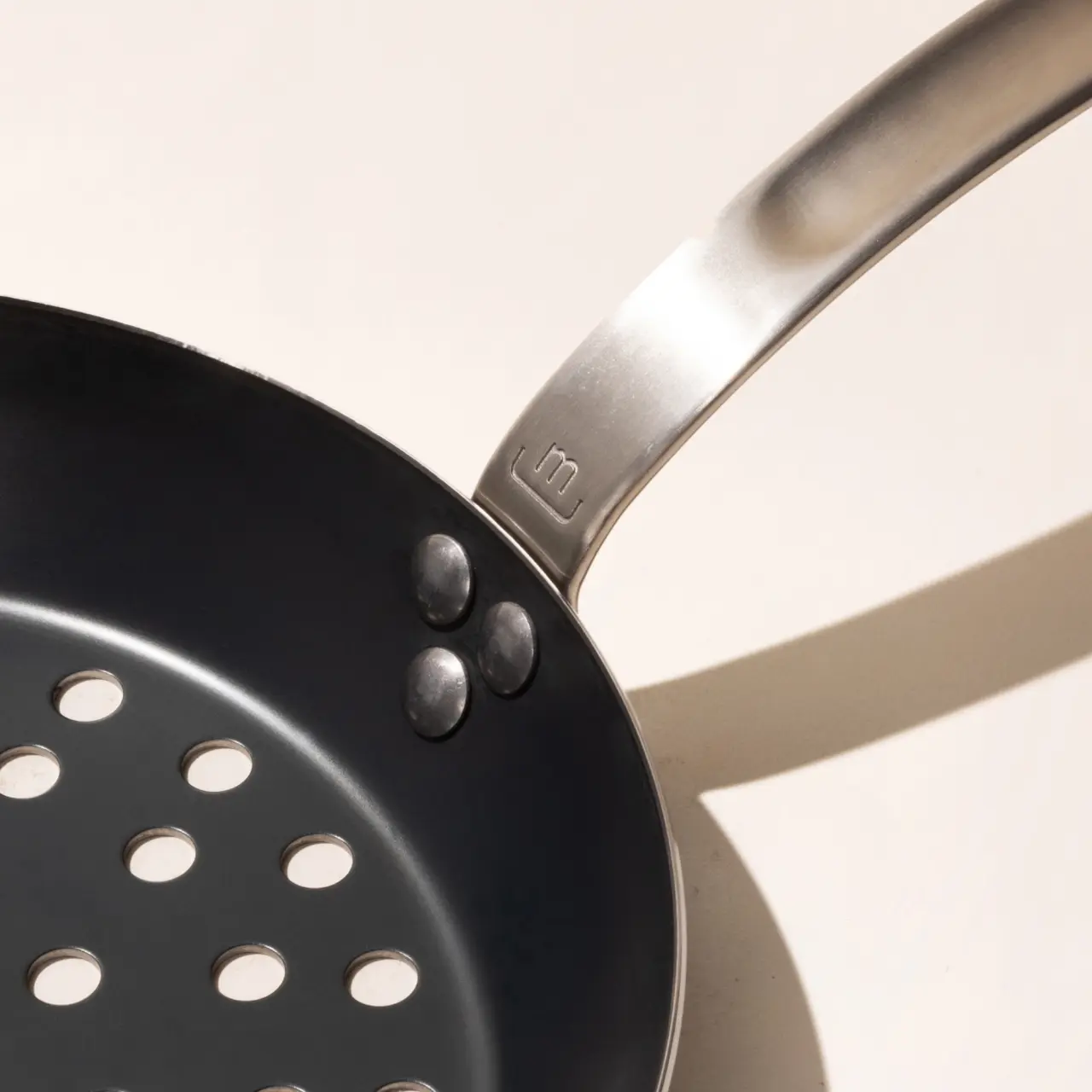 A stainless steel spatula with round cutouts rests on a non-stick frying pan with three small round objects that resemble chocolate candies on its surface, creating a minimalist composition with cast shadows.