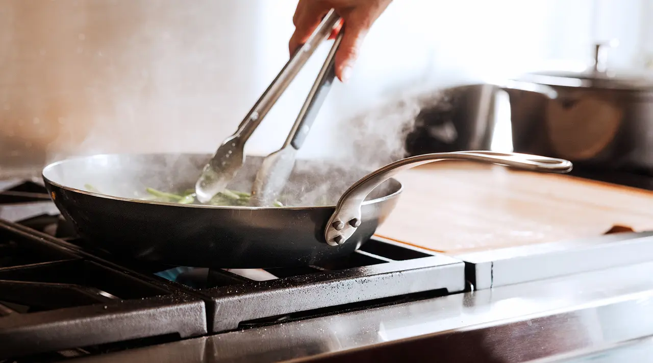 A person is sautéing food in a frying pan on a stove, with steam rising from the pan.