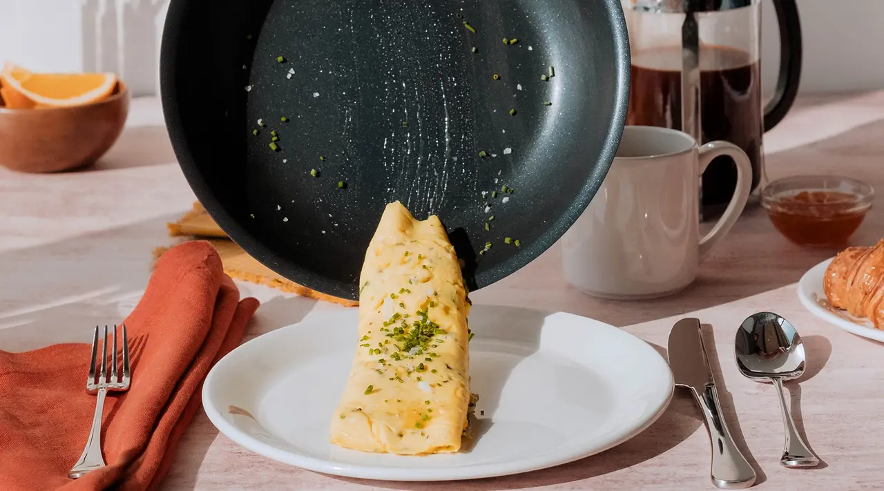 A crepe is being flipped onto a plate from a pan as part of a breakfast setting with utensils and beverages on the table.