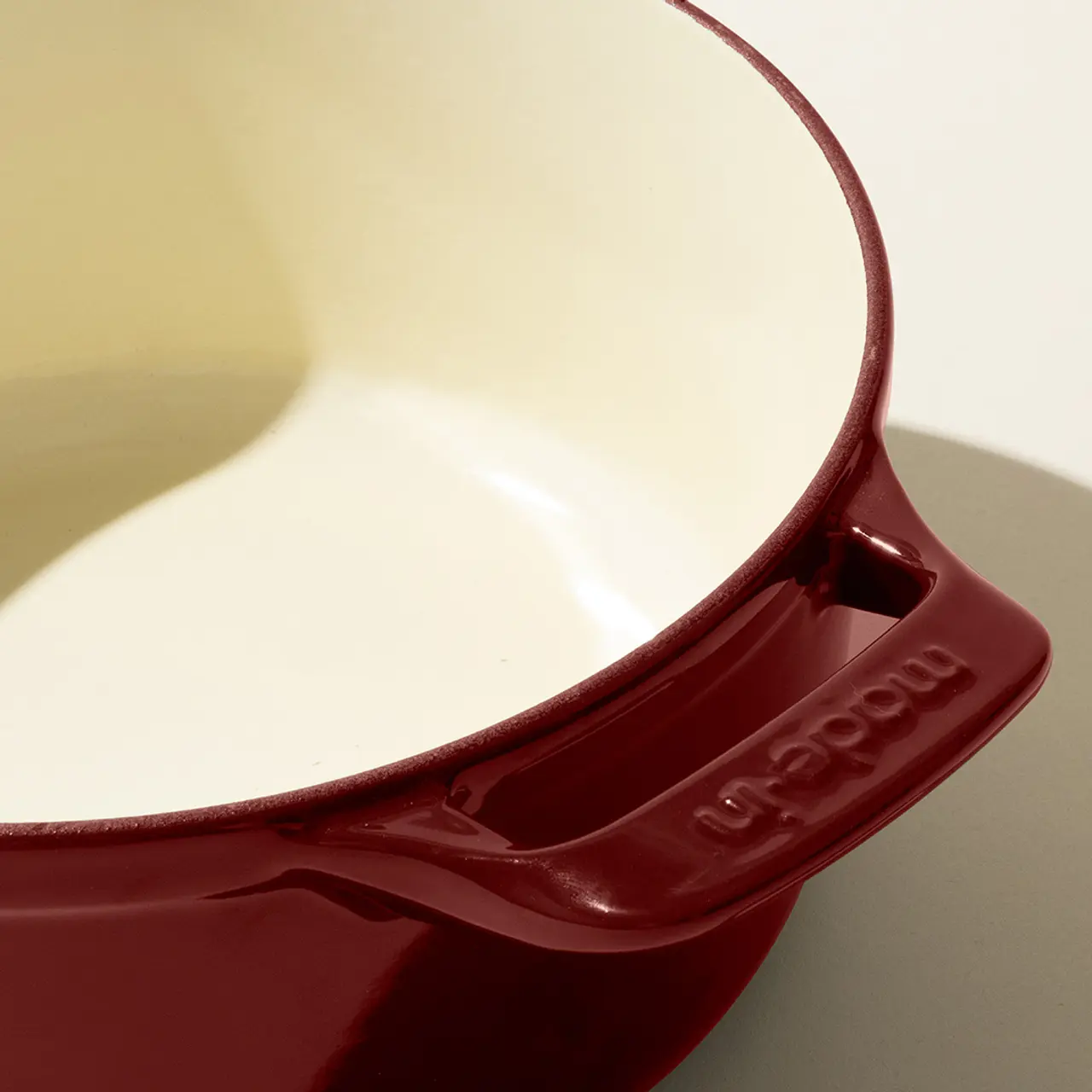 The image shows a close-up of a red enameled cast iron pot with a cream interior, focusing on the pouring spout with the brand name embossed.