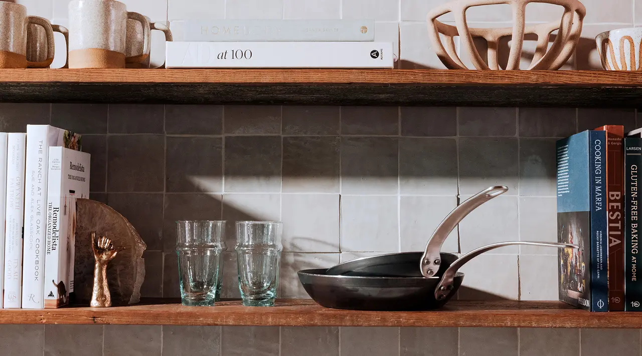 A well-organized kitchen shelf displays a collection of books, a frying pan, and various kitchenware against a tiled backsplash.
